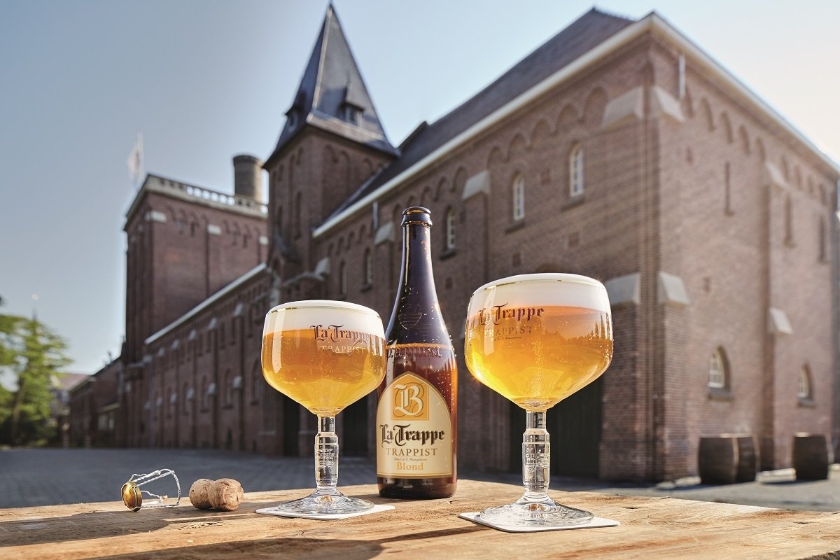 Botella y dos copas de La Trappe Blond con la fábrica de Koningshoeven de fondo