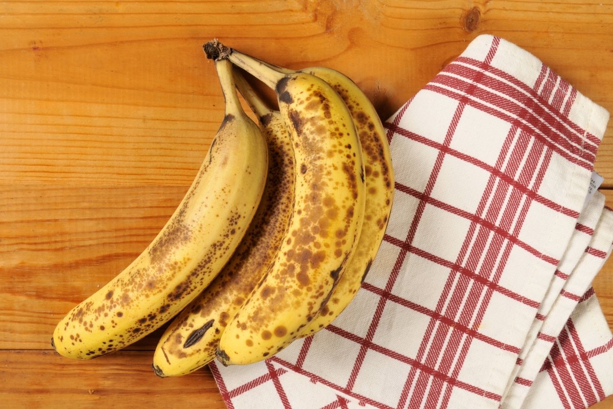 Cuatro plátanos muy maduros sobre una encimera con un paño de cocina blanco y rojo