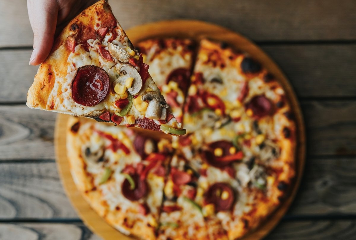 Una mano acerca una porción de pizza y de fondo se ve el resto de la pizza
