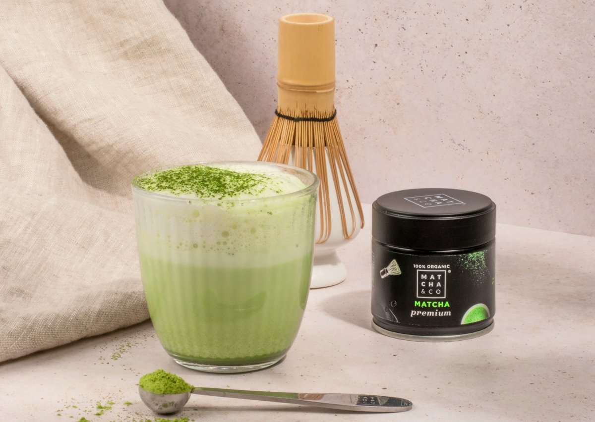 Vaso de matcha latte junto a un envase de matcha premium de Matcha & CO y los accesorios de la ceremonia del té matcha